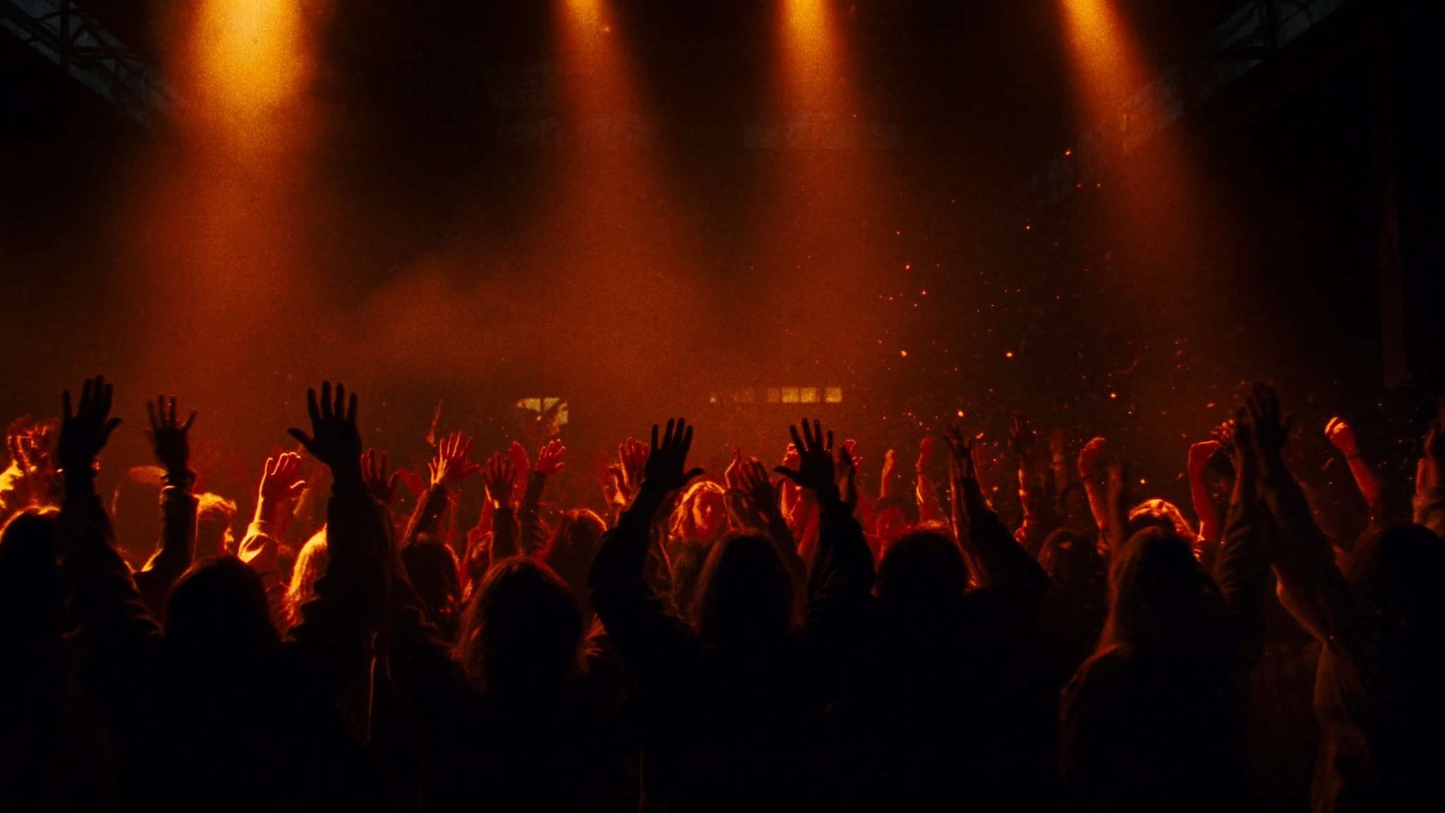 A dense crowd with arms raised, silhouetted in warm amber stage light and haze inside a dark warehouse venue