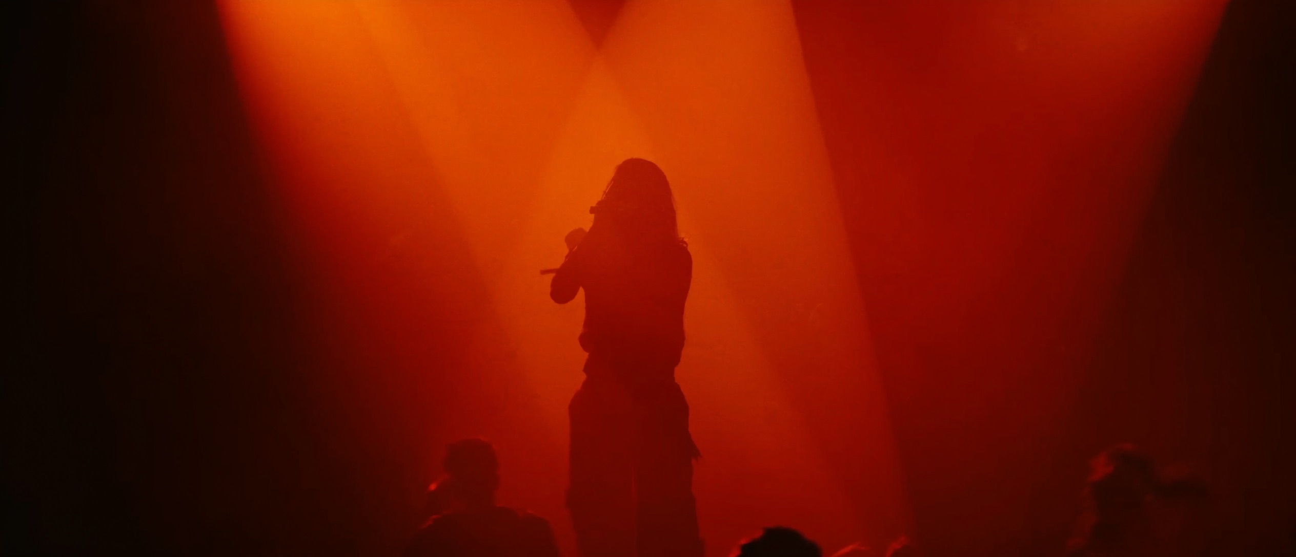 A lone figure silhouetted on stage under warm ember-orange spotlight beams, holding a microphone